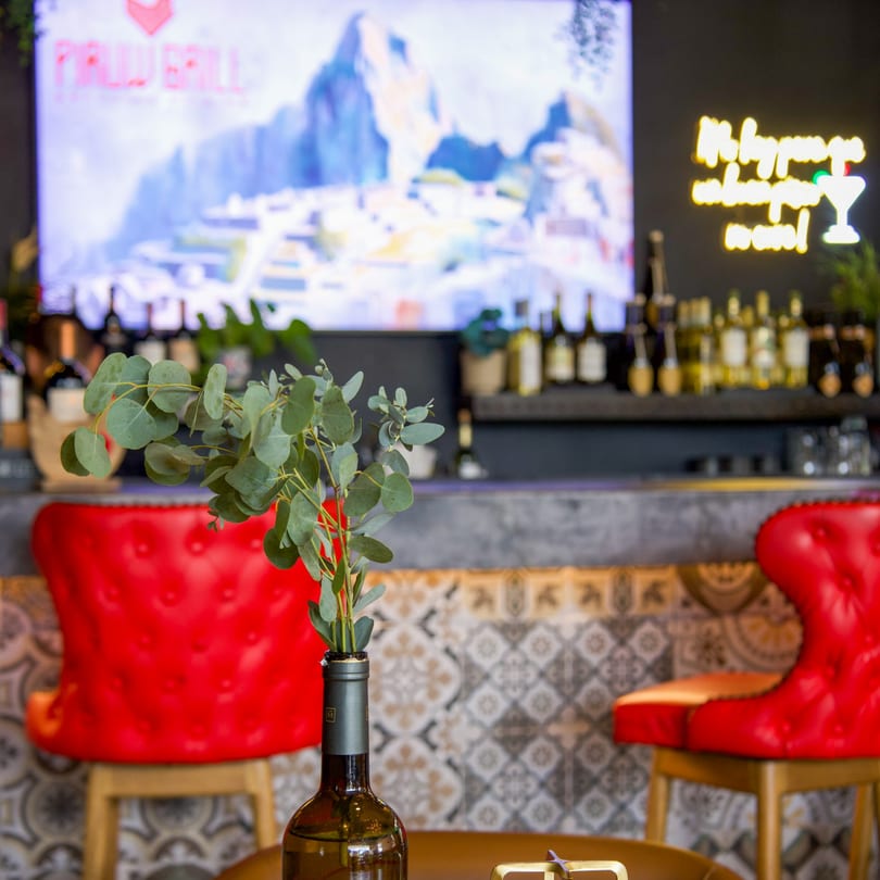 Eucalyptus in a wine bottle vase on a table with red bar stools and a bar in the background.