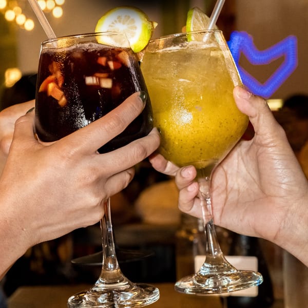 Two hands clinking glasses of dark red and yellow cocktails with fruit slices and straws.