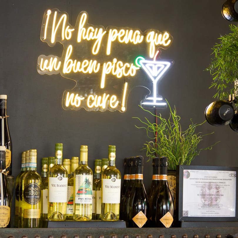 A neon sign with Spanish text and a martini glass above a display of wine and prosecco bottles.