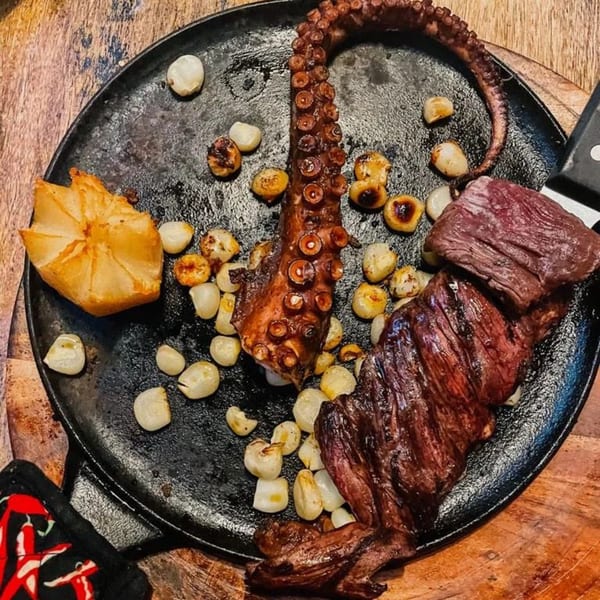 Grilled octopus tentacle, steak, roasted garlic, and a baked potato on a cast iron skillet.