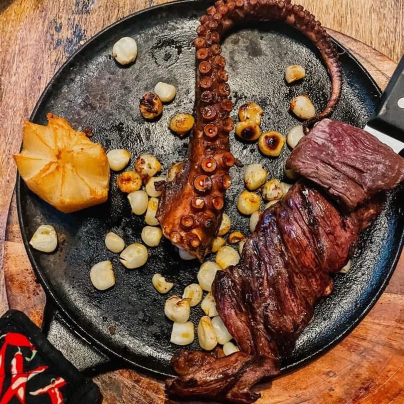 Grilled octopus tentacle, steak, roasted garlic, and a baked potato on a cast iron skillet.