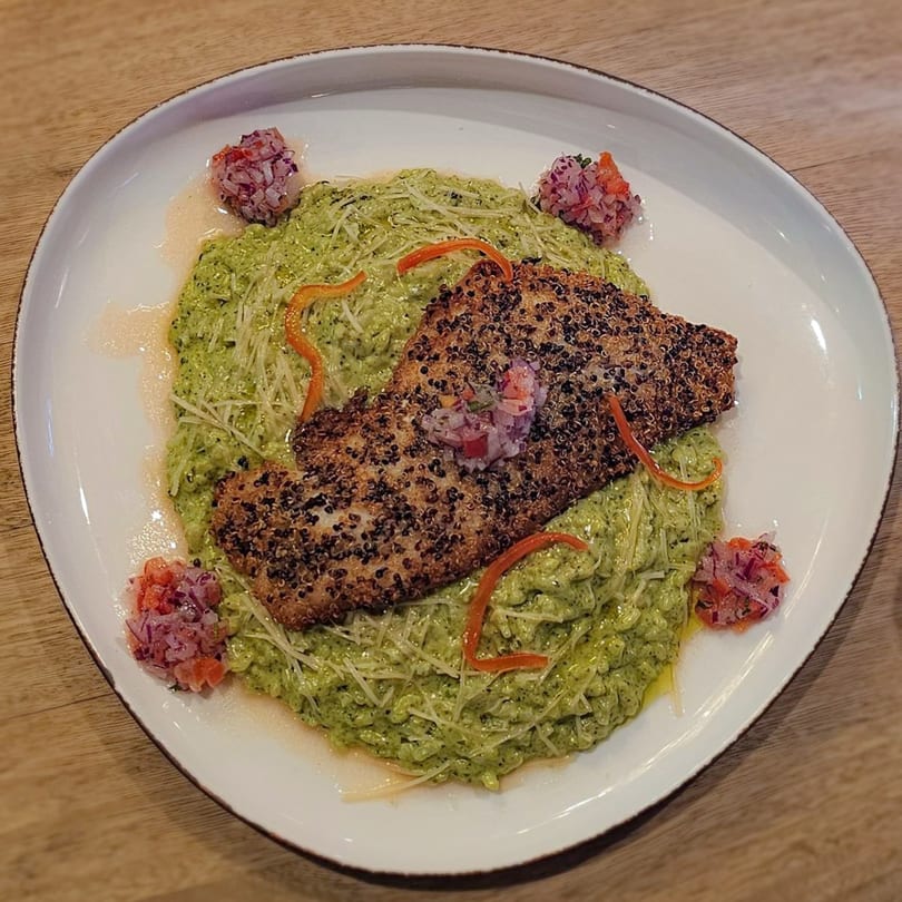 Quinoa-crusted fish over green risotto, garnished with red pepper strips and salsa on a white plate.