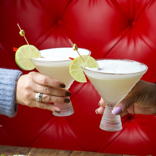 Two hands holding cocktails with lime wedges, toasting against a red tufted background.