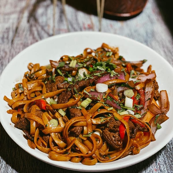 Close-up of a white plate filled with stir-fried noodles, beef, red onions, and green herbs.