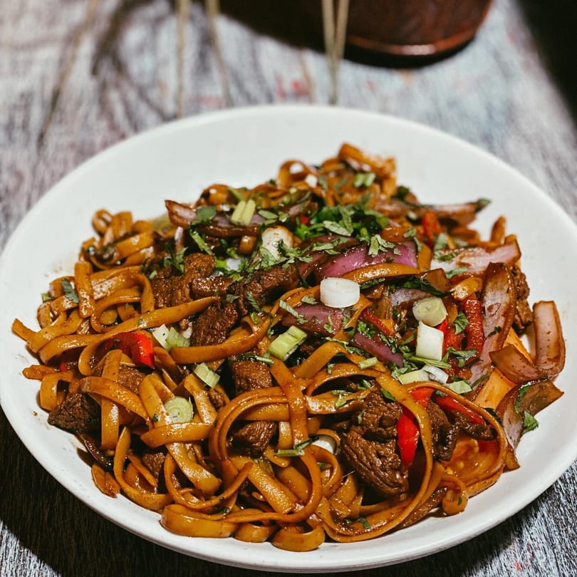 Close-up of a white plate filled with stir-fried noodles, beef, red onions, and green herbs.