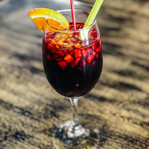 A glass of red sangria with diced fruit, an orange slice, and two straws on a wooden table.