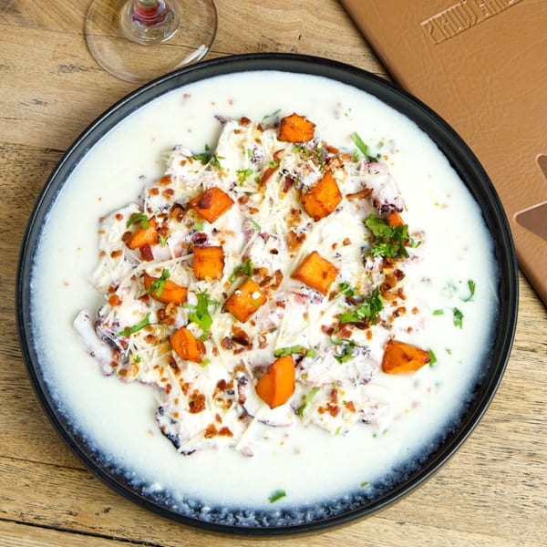 Creamy seafood dish with roasted butternut squash, bacon bits, and fresh herbs on a black plate.