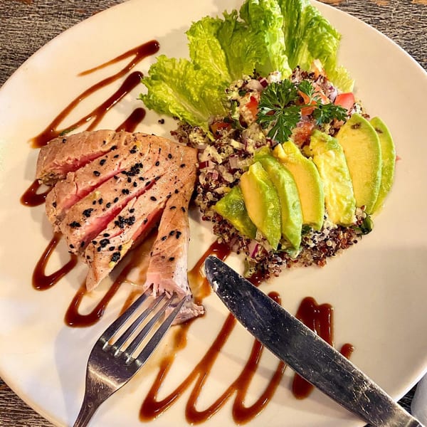 Seared tuna with sesame seeds, quinoa salad, avocado slices, and lettuce on a white plate.