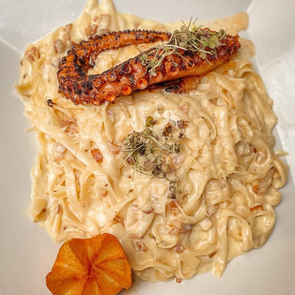 Creamy fettuccine pasta with a grilled octopus tentacle, garnished with microgreens and a plantain chip.